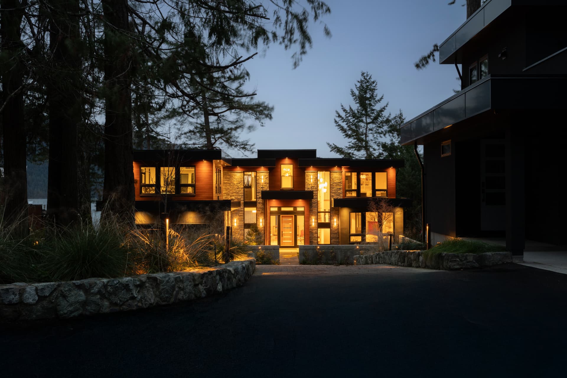 Stone and cedar residence glowing at blue hour behind mature firs